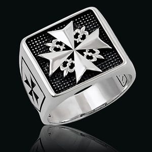 May include: A silver-colored ring with a square face featuring a Maltese cross design. The cross is set against a textured black background. The ring has a polished finish and a detailed design on the side.