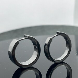 May include: A pair of dark metallic hoop earrings with a polished finish. Each earring features a hinged closure mechanism and is stamped with the hallmark 925 on the inner surface, indicating sterling silver construction with a dark plating.