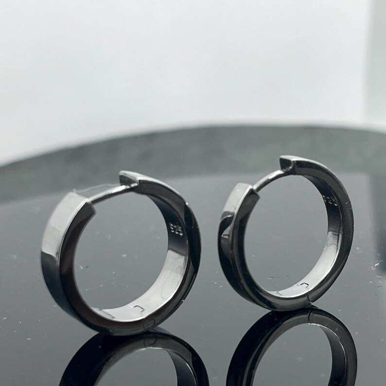 May include: A pair of dark metallic hoop earrings with a polished finish. Each earring features a hinged closure mechanism and is stamped with the hallmark 925 on the inner surface, indicating sterling silver construction with a dark plating.
