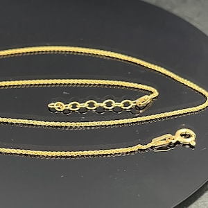 May include: A gold-colored wheat chain necklace featuring a delicate, braided link design. The necklace includes a small circular spring ring clasp at one end and a short extender chain with several larger circular links at the other end.