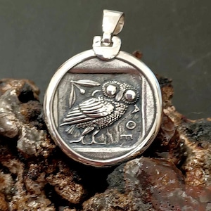 Può includere: Un ciondolo circolare in argento con un rilievo di un gufo di profilo, ispirato alle antiche monete greche. Il design include un ramoscello d'ulivo e le lettere AOE. Il ciondolo presenta una contromaglia in argento lucido sulla parte superiore.