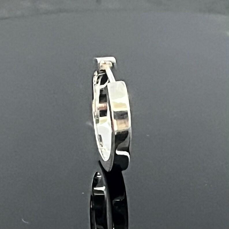 May include: A single silver-toned hoop earring with a polished, reflective surface. The earring features a hinged closure mechanism at the top, showing a smooth, rounded profile and a metallic finish that catches the light, creating a clean look.