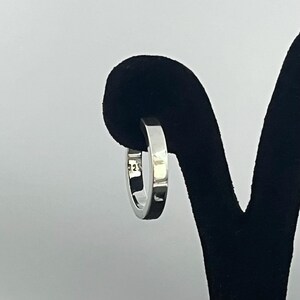 May include: A polished silver hoop earring with a flat, rectangular profile. The metal surface is smooth and reflective, featuring a clear 925 hallmark stamp on the inner band, indicating sterling silver composition for this circular jewelry piece.