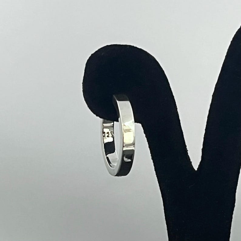 May include: A polished silver hoop earring with a flat, rectangular profile. The metal surface is smooth and reflective, featuring a clear 925 hallmark stamp on the inner band, indicating sterling silver composition for this circular jewelry piece.