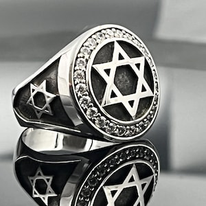 Może przedstawiać: Srebrny pierścionek z motywem Gwiazdy Dawida. Pierścionek ma dużą, okrągłą tarczę z wypukłą Gwiazdą Dawida, otoczoną pasmem małych, przezroczystych kamieni. Obręcz pierścionka jest również srebrna i ma Gwiazdę Dawida z boku.