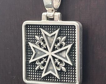 May include: A rectangular silver pendant featuring a raised Maltese cross design with fleur-de-lis accents at the center. The background of the pendant has a textured grid pattern, and it includes a polished silver bail at the top for hanging.