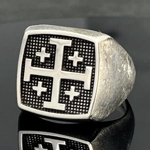 Könnte beinhalten: Ein silberfarbener Ring mit quadratischer Front und einem Jerusalemkreuz-Emblem. Das Kreuz ist poliert und hebt sich von einem dunklen, vertieften Hintergrund mit texturiertem Gittermuster ab. Die Ringschiene hat ein gebürstetes, mattes Finish.