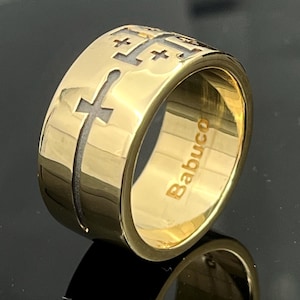 Könnte beinhalten: Ein breiter goldfarbener Bandring mit einem sich wiederholenden Muster aus eingravierten Jerusalemkreuzen mit vertieften dunklen Mitten. Die Innenseite des Rings ist poliert und zeigt deutlich den eingravierten Schriftzug Babuco in serifenloser Schrift.
