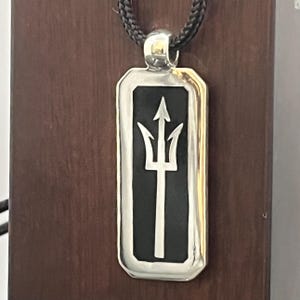 May include: Silver pendant necklace featuring a rectangular pendant with a black center and a trident design. The pendant is attached to a black cord. The necklace is displayed against a brown wooden background.