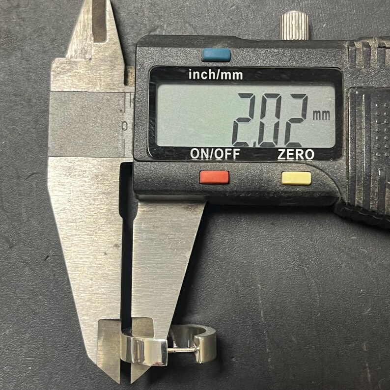 May include: A silver-colored hoop earring being measured by a black digital caliper. The digital display on the caliper clearly shows a measurement of 2.02 mm, indicating the thickness of the metal band of the earring in this close-up view.
