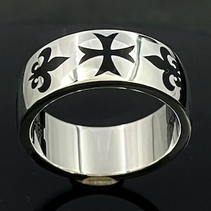 Könnte beinhalten: Ein breiter, silberfarbener Ring mit einem schwarzen Kreuz- und Fleur-de-Lis-Muster. Der Ring hat eine polierte Oberfläche und einen dunklen Rand. Das Design ist symmetrisch und kühn.