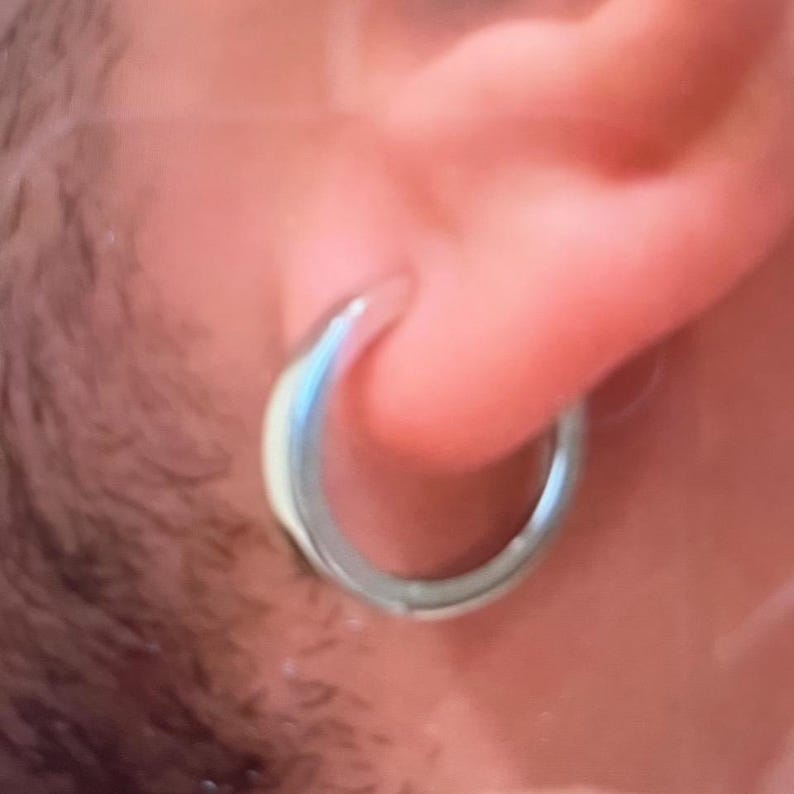 May include: A polished silver-toned hoop earring with a smooth, rounded surface. The circular metal band is designed to pass through an earlobe, featuring a reflective finish that catches the light. The earring has a simple, continuous loop structure.
