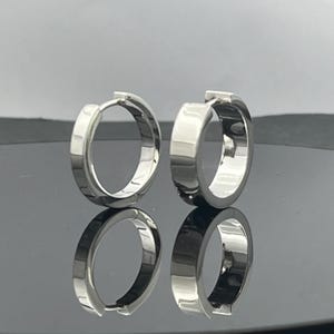 May include: A pair of polished silver-toned hoop earrings with a thick, flat-edged design. The earrings feature a hinged closure mechanism and exhibit a high-shine, reflective metallic surface, creating a clean and simple circular silhouette.