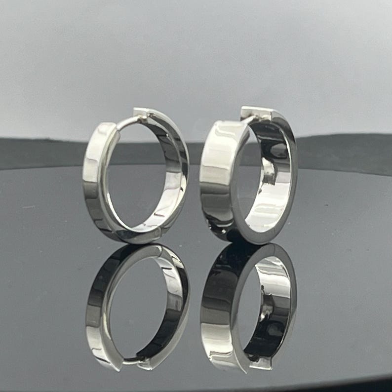 May include: A pair of polished silver-toned hoop earrings with a thick, flat-edged design. The earrings feature a hinged closure mechanism and exhibit a high-shine, reflective metallic surface, creating a clean and simple circular silhouette.