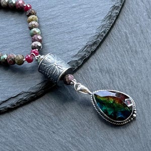 Könnte beinhalten: Eine Halskette mit einer Reihe facettierter, mehrfarbiger Perlen in Rot-, Grün- und Brauntönen. Die Kette enthält eine zylindrische, silberfarbene Metallperle mit eingraviertem Blumenmuster und einen tropfenförmigen Anhänger mit dunkler, irisierender Oberfläche.