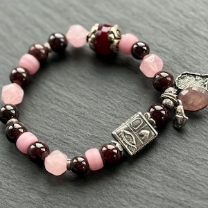 May include: A beaded bracelet featuring alternating dark red and light pink beads. The bracelet includes a silver charm with an oval pink stone and a decorative silver square. A stylish piece of jewelry.