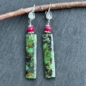 May include: A pair of dangle earrings featuring rectangular turquoise-colored stones with dark veining. Each earring has a small, round, red bead and a silver-colored leaf-shaped top. The earrings hang from silver-colored hooks.