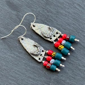 Op de afbeelding: Zilverkleurige oorhangers met getextureerde, bladvormige hangers. Elke oorbel heeft een cluster van rode, turquoise, gele en witte kralen. De oorbellen hebben een unieke, handgemaakte uitstraling.