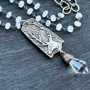 Op de afbeelding: Een zilverkleurige ketting met een rechthoekige hanger met een hertenontwerp. De hanger hangt aan een ketting van kleine, witte, afgeronde kralen. Onder de hanger hangt een helder, facetgeslepen kristal. De ketting ligt op een donkere, getextureerde ondergrond.