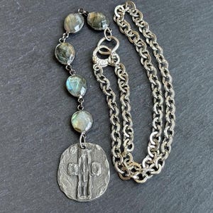 Op de afbeelding: Zilverkleurige ketting met een hanger met een textuurontwerp en een reeks ronde, iriserende edelstenen. De ketting heeft een dikke ketting en een decoratieve sluiting. De hanger heeft een uniek, reliëf ontwerp.