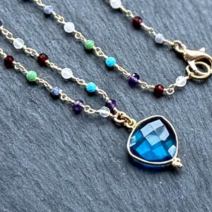 May include: A delicate gold necklace featuring a faceted, deep blue pendant. The necklace is adorned with small, colorful beads in various hues, including red, green, and purple. The clasp is gold-toned.