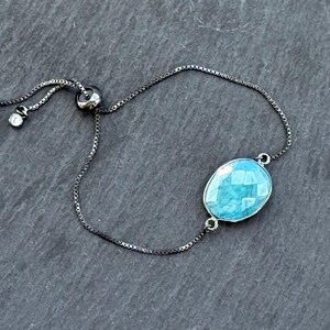 May include: A silver-toned adjustable bracelet with a faceted, oval-shaped, light blue gemstone. The bracelet features a delicate chain and a sliding bead for adjustment. The gemstone is set in a silver bezel.