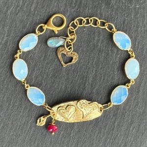 Könnte beinhalten: Ein goldfarbenes Armband mit ovalen, hellblauen Edelsteinen. Das Armband hat einen Herzanhänger, einen kleinen roten Edelstein und einen dekorativen goldenen Verschluss. Das Armband ist mit einer filigranen Kette versehen.