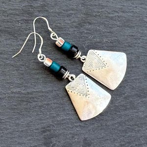 Puede incluir: Un par de pendientes plateados con un diseño geométrico. Cada pendiente presenta un gancho plateado, una sección de cuentas turquesas y negras, y un colgante plateado con un patrón de triángulo. Los pendientes se muestran sobre una superficie gris oscuro.