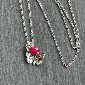 May include: A silver necklace featuring a butterfly pendant with a faceted, round, red gemstone. The butterfly pendant has intricate wing details. The necklace chain is delicate and silver.