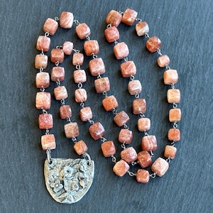 Op de afbeelding: Een ketting met vierkante, rood-oranje kralen geregen op een zilveren ketting. De ketting heeft een decoratieve zilveren hanger met bloemen- en bladerenontwerpen. De kralen zijn ongeveer 1,3 cm groot.