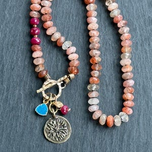 May include: A beaded necklace with a toggle clasp. The necklace features a mix of peach, pink, and red beads. A gold-colored charm with a flower design and a blue leaf charm hang from the clasp. The necklace is displayed on a dark gray surface.