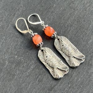 Op de afbeelding: Zilveren oorbellen met een visontwerp. Elke oorbel heeft een rechthoekige, getextureerde zilveren hanger met een vismotief, een oranje facetgeslepen kraal en een zilveren oorhaaksluiting. De oorbellen worden gepresenteerd op een donkergrijze ondergrond.