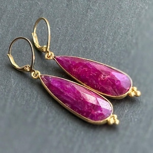 May include: A pair of drop earrings featuring faceted, teardrop-shaped magenta gemstones set in gold-toned metal frames. Each earring is suspended from a gold-toned lever-back closure, with small decorative metal beads at the base of each stone.