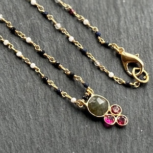 May include: A delicate gold necklace featuring a pendant with a large, oval-shaped green gemstone and three smaller, round pink gemstones. The chain is adorned with small white, black, and blue beads.