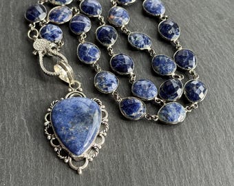 May include: A silver necklace featuring oval-shaped, faceted blue sodalite stones. The necklace includes a heart-shaped pendant with a matching blue stone, set in an ornate silver frame. The necklace is displayed on a dark surface.