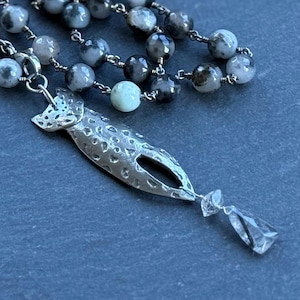 May include: A necklace featuring a chain of round, mottled grey, black, and cream-coloured beads. A textured, silver-toned metal pendant shaped like a stylised fish hangs from the chain, accented by a small, clear, faceted crystal drop at the bottom.