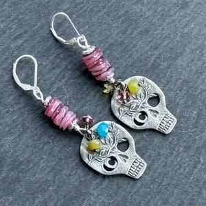 Op de afbeelding: Een paar zilveren schedeloorbellen met oorhaaksluitingen. Elke oorbel heeft een schedelhanger met bloemendetails, geaccentueerd met roze, blauwe en gele kralen. De oorbellen zijn geplaatst tegen een donkergrijze achtergrond.