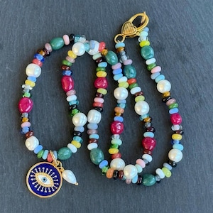 May include: A vibrant beaded necklace with a gold clasp and an evil eye charm. The necklace is made up of a variety of beads in shades of red, green, blue, yellow, and white. The pendant is a gold circle with a blue eye design.