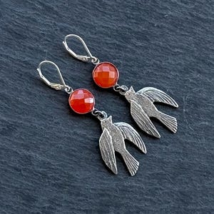 Op de afbeelding: Zilverkleurige oorbellen met een facetgeslepen, ronde, oranjerode edelsteen en een vogel silhouet. De oorbellen hebben oorhaaksluitingen. Gepresenteerd op een donkergrijze ondergrond.
