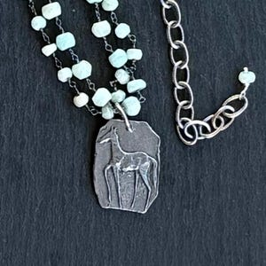 Op de afbeelding: Zilverkleurige ketting met een hanger met een herten silhouet. De ketting heeft een dubbele streng lichtblauwe edelsteen kralen en een kettingverlenger. De hanger is rechthoekig met een getextureerd oppervlak.