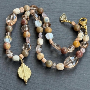 Op de afbeelding: Een ketting met een mix van bruine, beige en transparante kralen. De ketting heeft ronde, ovale en facetgeslepen kralen, een goudkleurige hanger en een sluiting. De kralen zijn geregen op een koord. Ongeveer 45 cm lang.