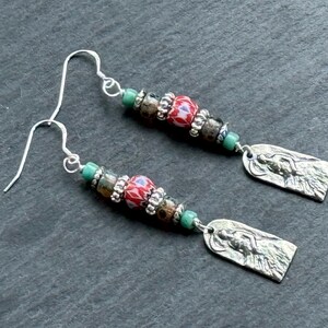 Op de afbeelding: Een paar oorhangers met zilveren haken. Elke oorbel heeft een combinatie van kleurrijke kralen, waaronder rood, groen en bruin, en een zilveren rechthoekige bedel met een gedetailleerde figuur.