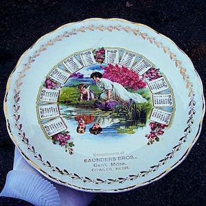 May include: Vintage decorative plate with a scalloped edge and gold trim. The center features a calendar design with a pastoral scene of a woman and child by a pond. The plate has the text "Compliments of SAUNDERS BROS., Gen'l. Mdse., COWLES, NEBR."