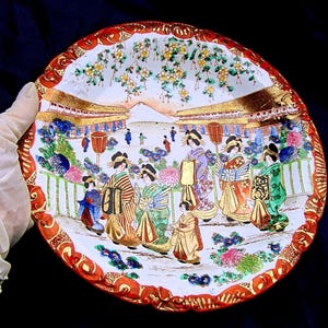 May include: A decorative plate with a Japanese scene. The plate features a detailed illustration of figures in traditional attire, buildings, and floral elements. The rim is red with gold accents, and the overall color palette is vibrant.