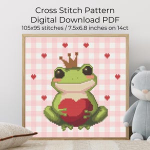 May include: Cross stitch pattern digital download PDF. The framed artwork features a green frog wearing a crown and holding a red heart. The background is a pink and white gingham pattern with red hearts. The text reads: 105x95 stitches / 7.5x6.8 inches on 14ct.