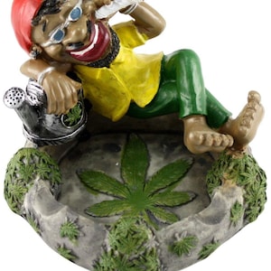 May include: A ceramic ashtray featuring a cartoon figure with a red hat, sunglasses, and a yellow shirt, sitting on a green leaf with a marijuana leaf design. The ashtray is decorated with green leaves.