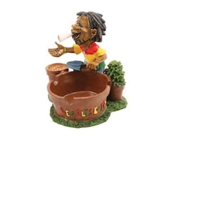 May include: A brown ceramic ashtray with a cartoon figure of a person smoking a cigarette. The ashtray has the words "Legalise It" written on it.