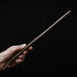 May include: A handcrafted wooden wand with a textured, natural finish. The wand is held against a black background, showcasing its rustic appearance. The wand is approximately 30 cm long.