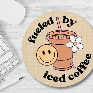 Puede incluir: Una alfombrilla de ratón redonda de color marrón con el texto "Fueled by iced coffee" en blanco y negro. Una cara sonriente de dibujos animados y una taza de café helado de dibujos animados con una pajita rosa y una flor blanca están en la alfombrilla de ratón.