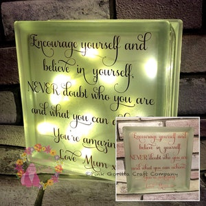 Op de afbeelding: Een lichtgevend glazen blok met de tekst "Encourage yourself and believe in yourself. NEVER doubt who you are and what you can achieve. You're amazing. Love Mum x" in zwarte letters. Het blok wordt verlicht met witte lampjes.
